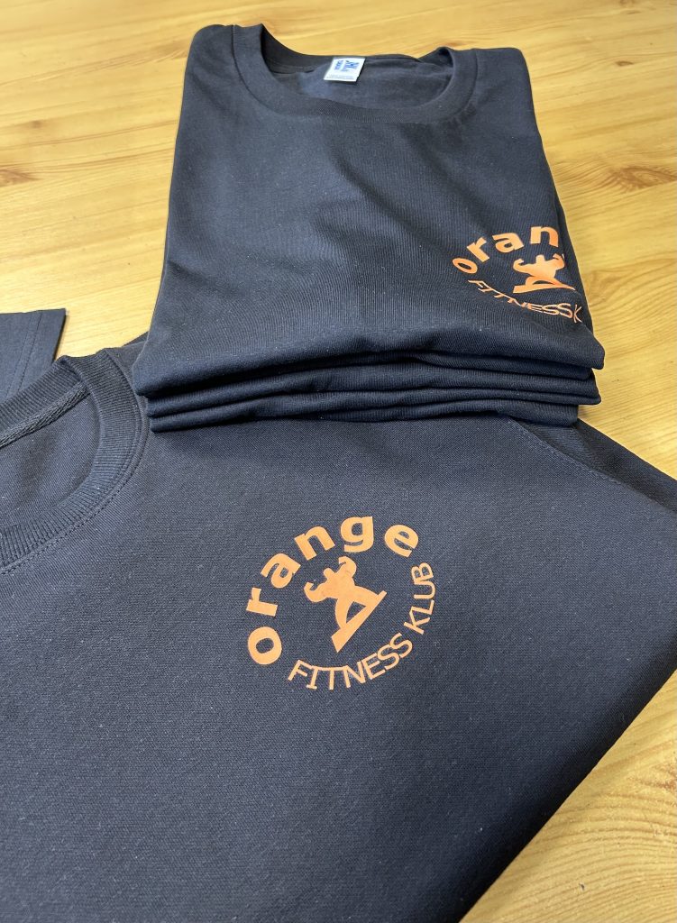 Stos czarnych koszulek i bluza z pomarańczowym logo Orange Fitness Klub, realizacja JustWearIt.pl, Środa Wielkopolska